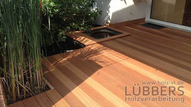 Terrasse mit Holzplanken, umgeben von Pflanzen und einem kleinen Wasserbecken in der Ecke.