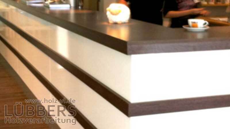 Theke mit dunkler Holzoberfläche und hellen Paneelen; im Hintergrund sind Mitarbeiter in einem Café aktiv.