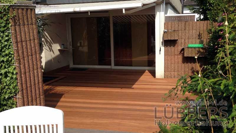 Terrasse mit Holzbelag, Blick auf große Fenster des Wohnzimmers und angrenzende grüne Pflanzen.